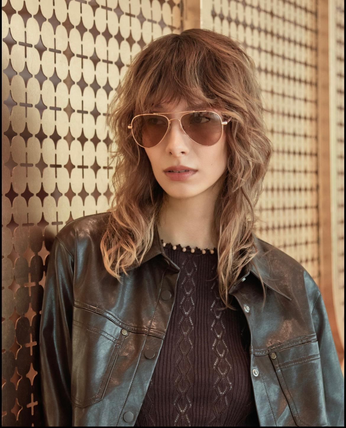 Editorial portrait of a model wearing aviator sunglasses, a dark knit top and a leather jacket against a patterned wall