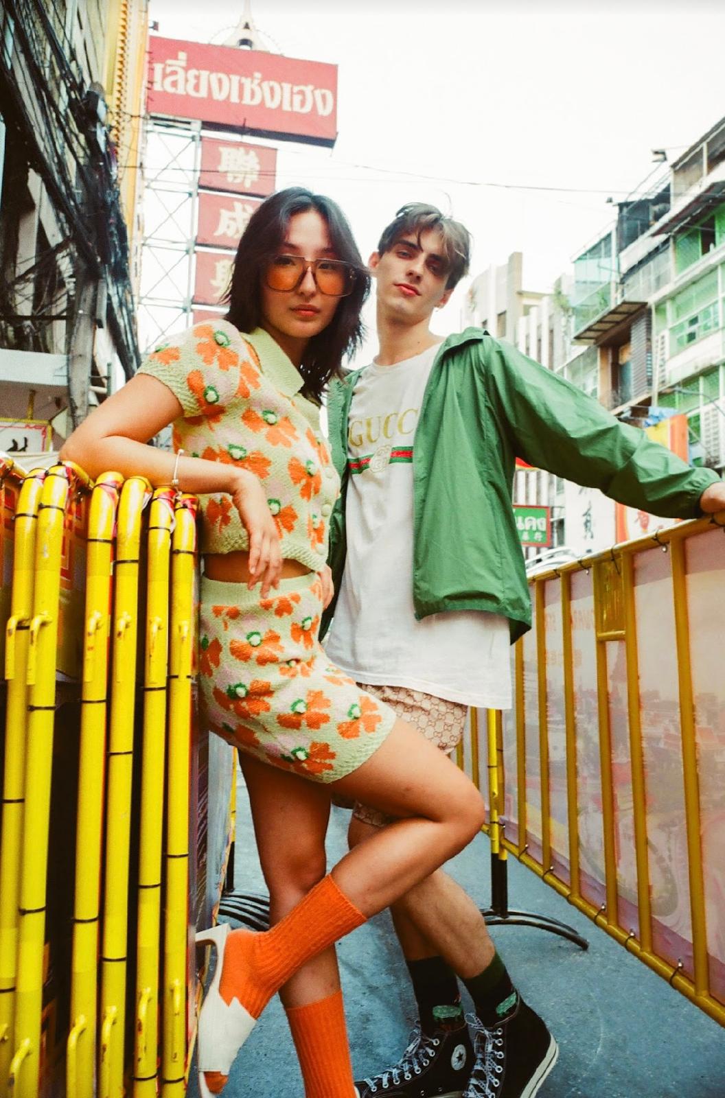 Two editorial models in floral orange knitwear and a green jacket beside yellow street barriers